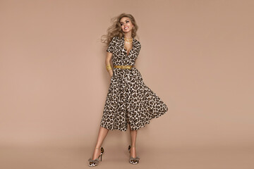 Stylish female model wearing a fashionable leopard-print dress is posing on a beige background in the studio.