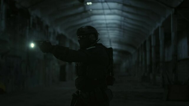 Special forces operator in stealthy black gear drawing his glock17 handgun from the holster and aiming with the flashlight turned on in a dark abandoned warehouse