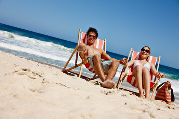 Fototapeta premium Couple, sunglasses and relax at beach for travel, bonding and tropical holiday with chair on sand. Man, woman and ocean outdoor for trip, love and vacation with sunshine, smile and weekend in Greece