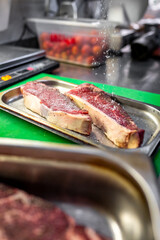 A close-up of a seasoned steak on a metal tray in a professional kitchen, with salt being sprinkled on top. The blurred background highlights the cooking process, emphasizing the preparation stage. 