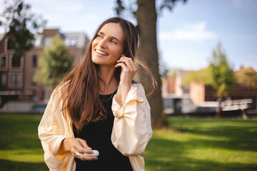 Brunette woman puts wireless headphones into her ears. Female hands hold two white glossy wireless Bluetooth headphones. Girl hold case and put on earphone. Happy laughing girl listen music, audiobook