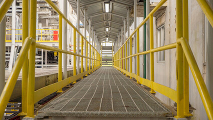 Yellow Handrail Walkway in Industrial Setting. A yellow handrail walkway leads into a industrial area.  The walkway is made of metal and has a grid pattern.
