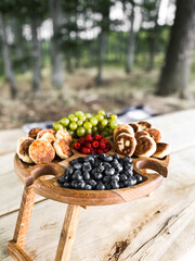 A wooden plate with fruits and cheeses on the background of a field with sheep. The concept of a picnic in nature