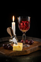 A glass of red wine with a bottle, a piece of cheese and grapes on an old wine barrel and a candle