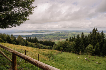 Scenic view of rolling hills, forests, and lakes in Bavaria, framed by a wooden fence and cloudy skies above the lush green landscape..