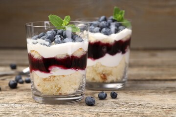 Tasty trifle dessert. Sponge cake, blueberries, jam and whipped cream in glasses on wooden table, closeup