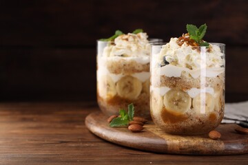 Tasty trifle dessert. Sponge cake, banana and whipped cream in glasses on wooden table, space for text
