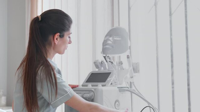 Female cosmetologist using modern hydrafacial machine in clinic. Young Caucasian professional is setting equipment as she touches screen while preparing for treatment at salon. 