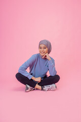 A cheerful girl wearing a colorful hijab is sitting casually against a vibrant pink backdrop