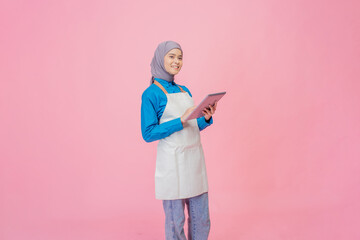 Fototapeta premium A joyful young woman in a stylish apron holds a tablet, smiling against a vibrant pink backdrop