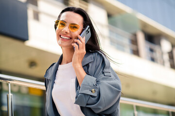 Photo of cheerful pretty lady dressed grey jacket dark eyewear smiling communicating modern device outdoors town street