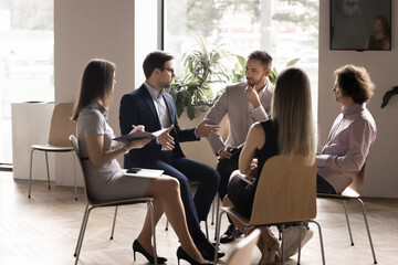 Serious boss instructing team of employees sitting in circle in office interior. Mentor, coach teaching group of interns. Employees listening to business project leader, writing notes on meeting