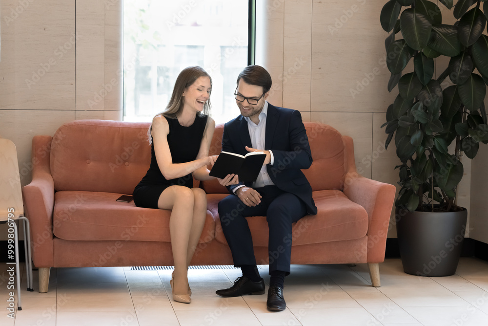 Poster two cheerful couple of colleagues discussing project in office lobby, sitting on couch, reading note