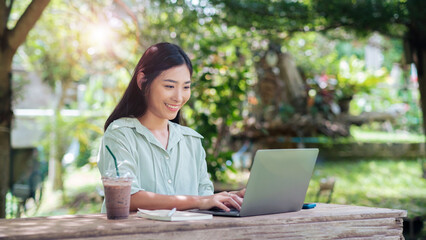 Asian business woman work outside enjoying fresh air working laptop computer portable device wireless remote digital technology, outdoor nature with drink, sitting park cafe under tree with sunlight