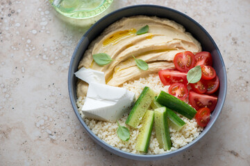 Hummus bowl with couscous, feta cheese and fresh vegetables, horizontal shot on a beige granite background, elevated view