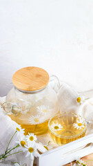 Glass teapot, cup, chamomile tea, daisies on white napkin on wooden box tray on light background.