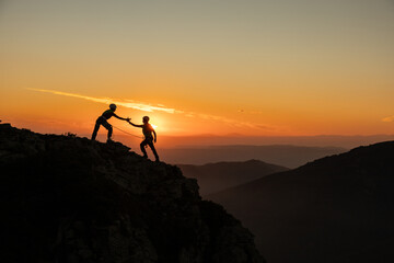 Two people are climbing a mountain together, one of them is helping the other. The sun is setting in the background, creating a beautiful and serene atmosphere