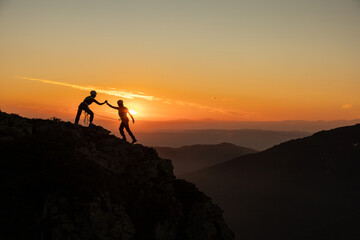 Two people are climbing a mountain together, one of them is holding the other's hand. The sun is setting in the background, creating a warm and peaceful atmosphere