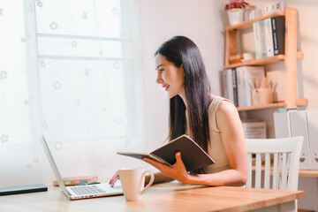 Cheerful asian woman sits at home office working on laptop and holds notebook and enjoys a cup of coffee, surrounded by a bright and airy atmosphere.