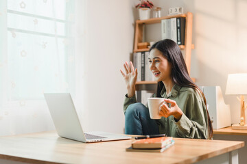 Asian woman sitting at wooden table smiling and waving while holding cup engaged in video call on her laptop. cozy room features natural light and bookshelf.