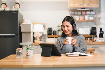 Young woman sits comfortably in her kitchen sipping coffee while using her tablet. She enjoys a...