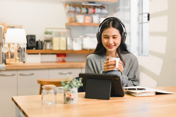 Young asian woman wearing headphones enjoys warm drink while using tablet in cozy kitchen setting....