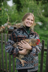 Pretty blonde farmer holding chicken in hands while standing in yard in rural area.