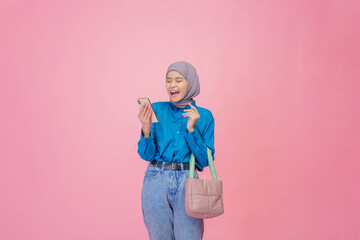 A young woman in a stylish hijab is happily using her smartphone against a vibrant pink backdrop