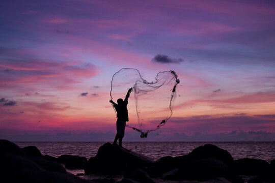 Silhouette of a fisherman standing on rocks and casting a fishing net at dusk, Thailand