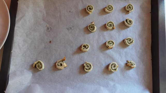 Hand with tongs picking up warmed cinnamon roll pieces one by one from parchment paper on a baking tray. The rolls are arranged evenly, showing their spiral pattern