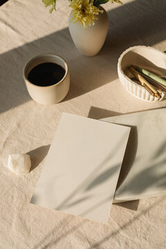 Overhead view of a blank piece of card on a table with assorted stationery, a cup of black coffee and a vase with flowers in sunlight
