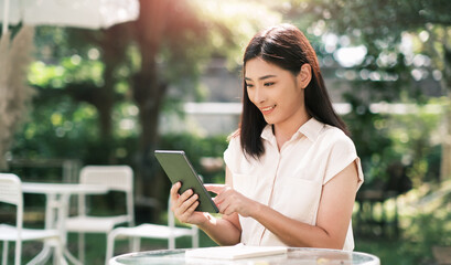 Asian business woman working outside freelancer using digital tablet device wireless remote communication technology, connectivity online worker casually enjoying outdoor in nature at cafe sunlight.