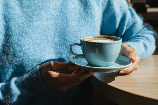 Close-up of a woman in a blue jumper sitting at a table holding a cappuccino in a blue cup