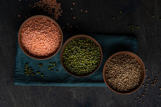 Overhead view of three bowls with red lentils, coriander seeds and green mung beans on a folded napkin on a wooden table