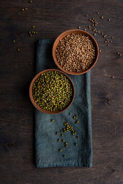 Overhead of bowls of coriander seeds and green mung beans on a folded napkin on a wooden table