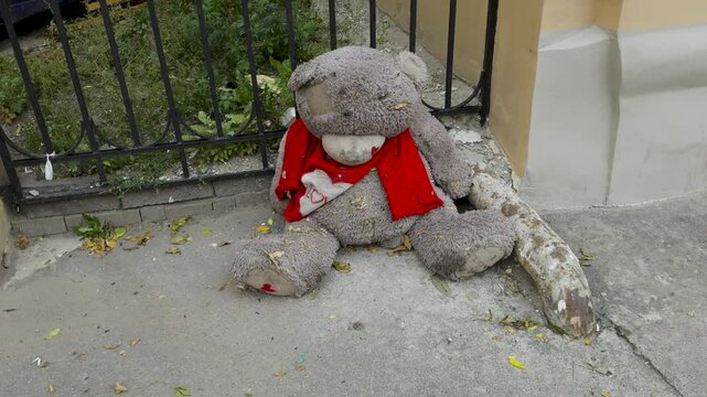 A large, worn teddy bear wearing a red scarf sits abandoned by a metal fence in Saint Petersburg. The bear shows signs of wear, with patches and dirt covering its surface, creating a melancholic