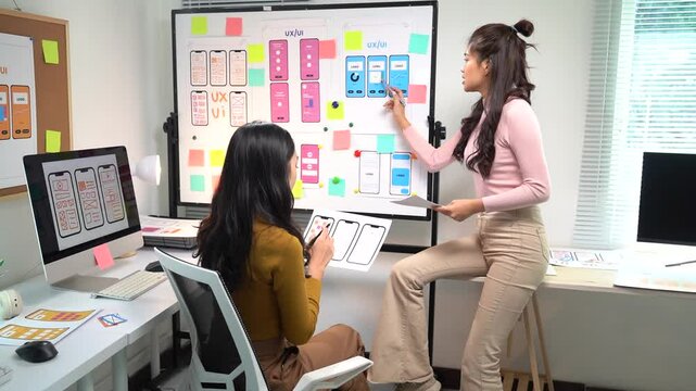 Two asian women designers are discussing a ux ui design for a mobile application, using a whiteboard and sticky notes in a modern office