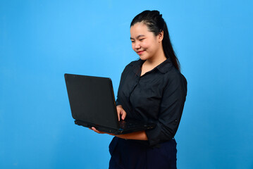 joyful asian woman in black shirt standing holding laptop on blue backround smiling to camera