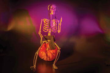 Skeleton figurine sitting on pumpkin, illuminated by mix of purple and orange lighting, creating...