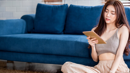 young beautiful Asian woman sitting on blue sofa and reading web by tablet at living room in the morning