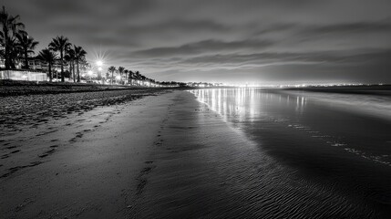 A black and white photo of a beach at night. The beach is empty and the water is calm. The sky is cloudy and the beach is lit up by the lights of the houses in the background