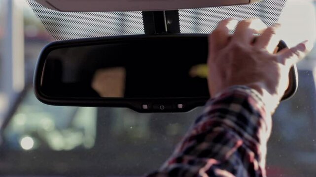 Close-up of a hand adjusting the rearview mirror inside a car, with the windshield and street scene in soft focus in the background