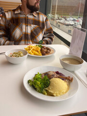 Millennial man in a plaid shirt sits at a white table with a hearty meal in front of him, featuring mashed potatoes, broccoli and big portion of protein. Cozy restaurant ambiance and tasty dinner