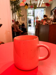 Mug with hot winter drink on a table in cozy cafe with bright red light. Blurred figures, plants, and festive Christmas decorations create warm and inviting atmosphere. Coffee shop environment with