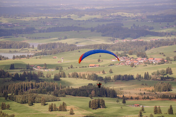 A paraglider soars over a scenic, rural landscape with rolling green hills, farms, and scattered...