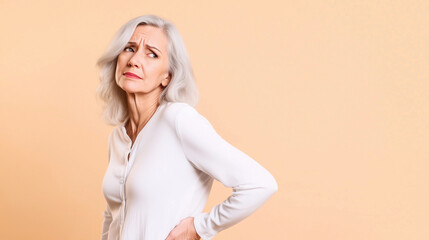 Naklejka premium A senior woman in white, leaning forward slightly as she clutches her lower back, her face etched with pain from spine issues, set against a neutral backdrop with room for messagin