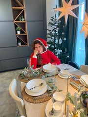 Millennial woman in a Santa suit sits thoughtfully and sad at a festive dining table, surrounded by Christmas decorations and a lit tree. Moment of holiday reflection and problem loneliness in