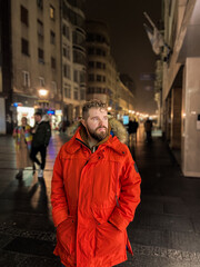 Man walking in a bustling nighttime city street, wearing a bright orange parka. Serene contemplation against the backdrop of lively city lights at night. Urban life concept