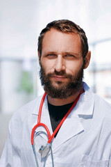 Confident bearded male doctor wearing a white lab coat with a stethoscope around his neck stands in a bright modern Israeli medical facility during daylight hours. Family doctor patients consultation