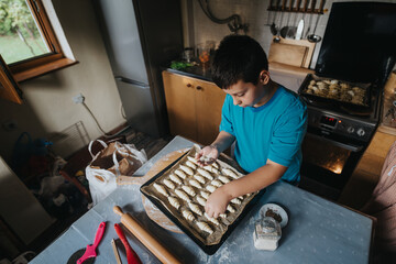 A young boy enthusiastically prepares homemade pastries on a baking tray in a cozy kitchen,...